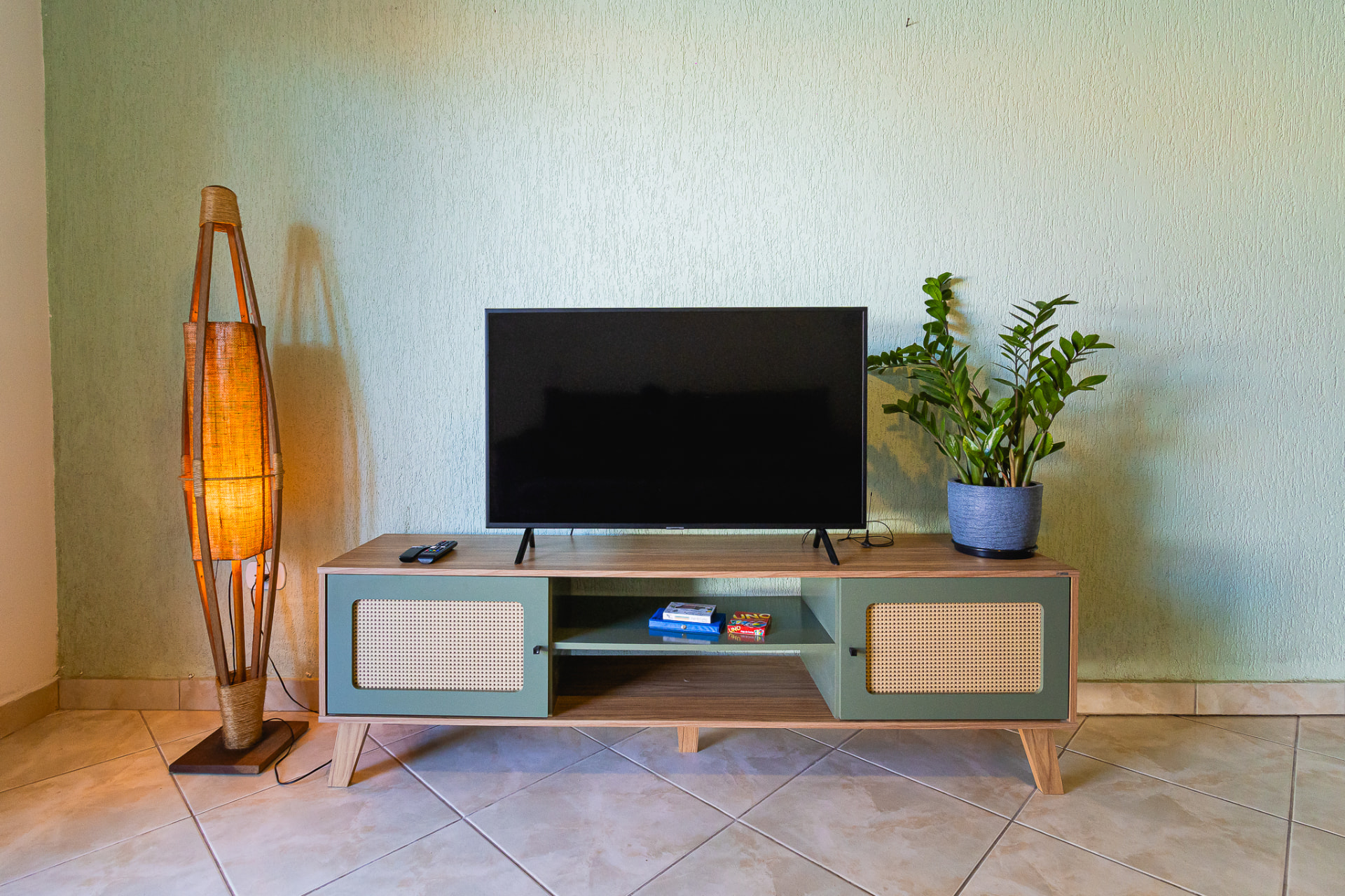 Sala de estar com rack de madeira e palhinha, TV, luminária artesanal e planta