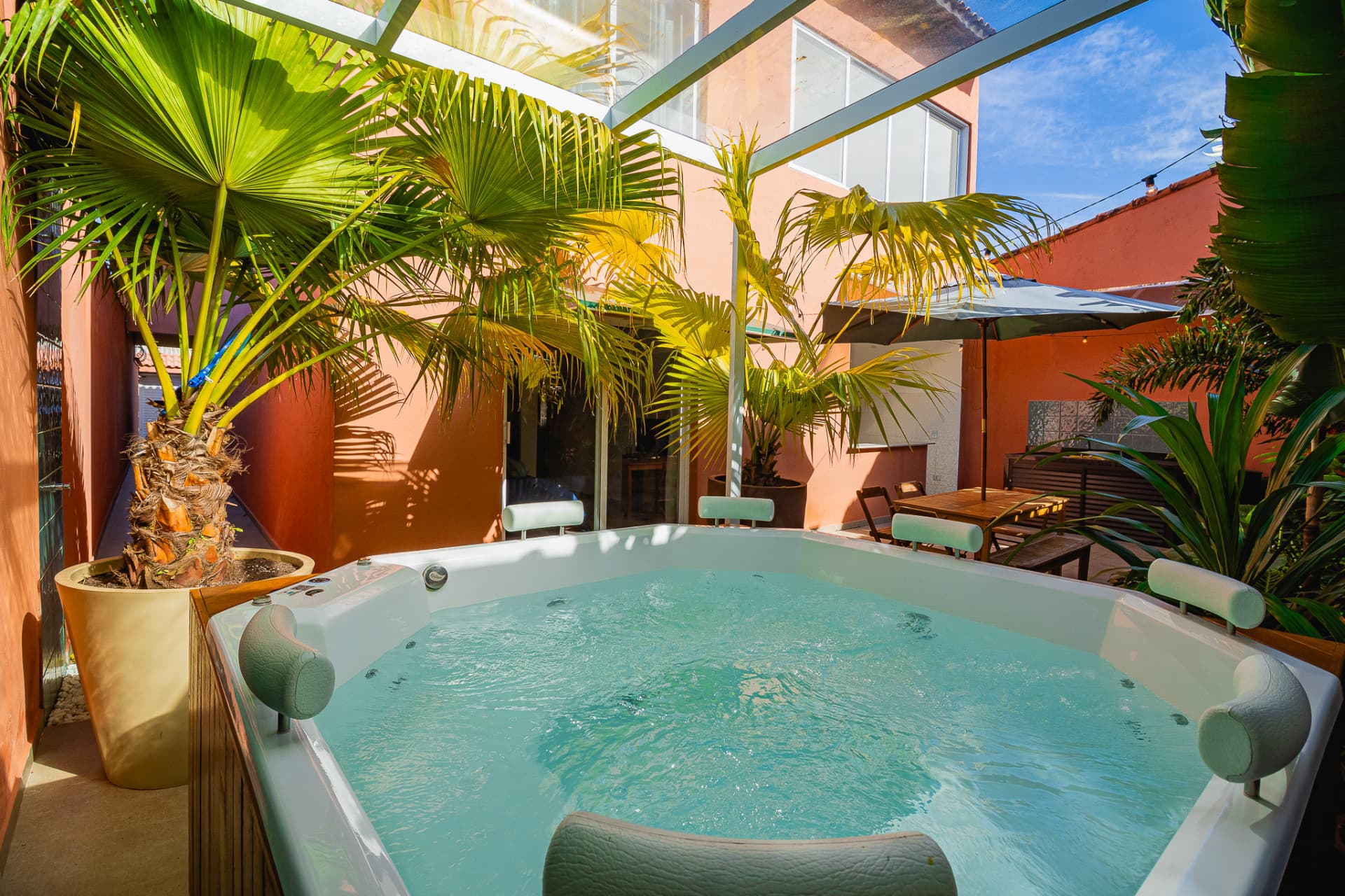 Jacuzzi ao ar livre em área externa com paredes terracota, palmeiras e mesa sob guarda-sol